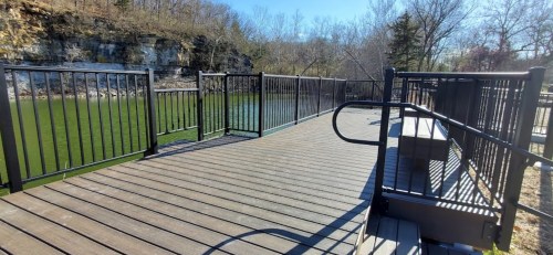 A wooden deck with black railings overlooking a calm green river, surrounded by trees and rocky cliffs.