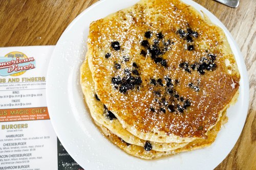A stack of golden pancakes with blueberries, dusted with powdered sugar, next to a diner menu.