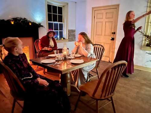 Four women in period clothing gather around a candlelit table in a cozy, warmly lit room with a fireplace.
