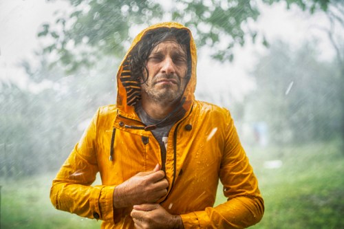 A man in a yellow raincoat stands in heavy rain, looking pensive with a blurred, rainy background.