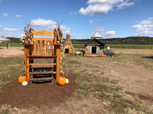A wooden pumpkin patch sign with pumpkins nearby, set against a blue sky and a rustic barn in the background.