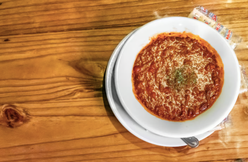 A bowl of red sauce with herbs on a wooden table, accompanied by a spoon and two sugar packets.