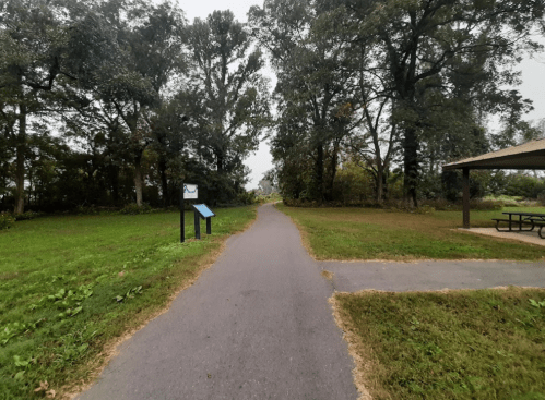 A paved path through a grassy area, lined with trees, leading to a picnic shelter and informational signs.