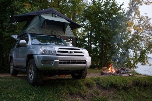 A silver SUV with a rooftop tent parked near a campfire by a lake, surrounded by trees.