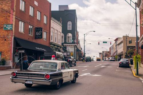 A vintage police car drives down a city street lined with shops and buildings under a cloudy sky.