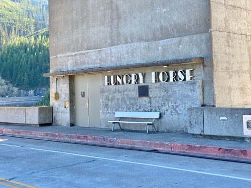 Concrete building with the words "HUNGRY HORSE" on the wall, a bench in front, surrounded by trees and mountains.