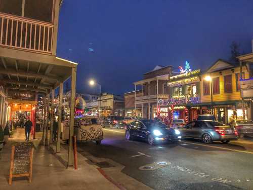 A lively street scene at dusk, featuring shops adorned with lights and cars passing by in a festive atmosphere.