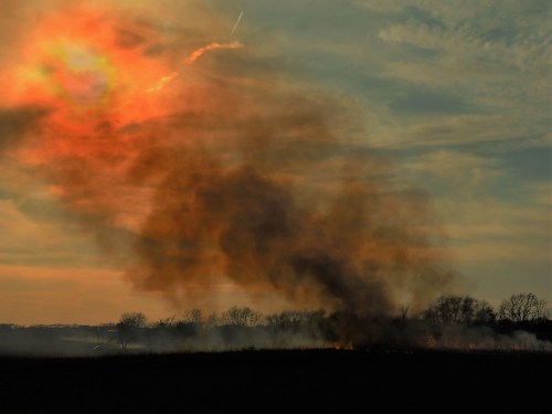 A smoky landscape at sunset, with dark smoke rising against a colorful sky.