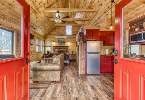 Cozy interior of a wooden cabin with red doors, featuring a living area, kitchen, and large windows for natural light.
