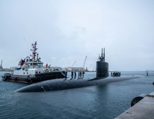 A submarine docked at a harbor with crew members on board, while a tugboat assists nearby in overcast weather.