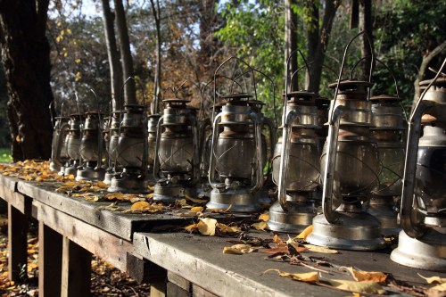 A row of vintage lanterns on a wooden table, surrounded by autumn leaves and trees in a sunny outdoor setting.