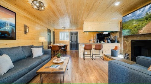Cozy cabin interior with wooden walls, a living area, kitchen bar, and dining space, featuring modern decor and a fireplace.