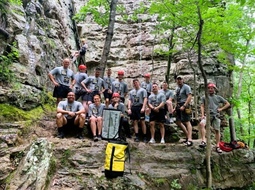 A group of climbers in matching shirts and helmets poses at a rock climbing site surrounded by trees.