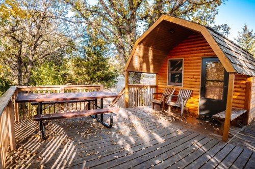 Cozy wooden cabin with a deck, picnic table, and chairs, surrounded by trees and autumn foliage.
