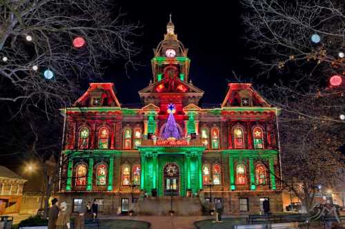 A historic building illuminated with festive red and green lights, featuring a large Christmas tree decoration.