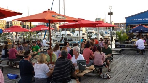 A lively outdoor dining area with people at picnic tables under red umbrellas, overlooking a marina at sunset.