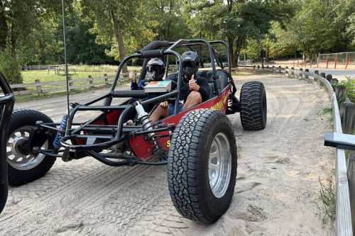 A dune buggy parked on a sandy path, with two people inside giving thumbs up, surrounded by trees.