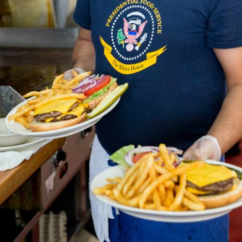 A server in a Presidential Food Service shirt holds two plates of burgers with toppings in a kitchen setting.