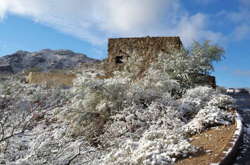 A stone building surrounded by snow-covered shrubs and mountains under a partly cloudy sky.