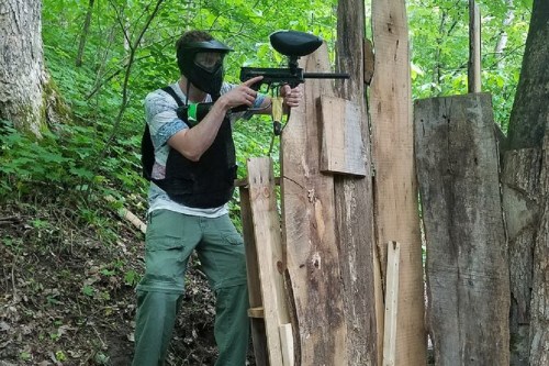 A person in a mask and vest aims a paintball gun from behind wooden barriers in a wooded area.