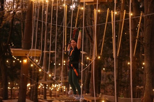 A child in a harness smiles while navigating a rope course, surrounded by trees and twinkling lights at dusk.