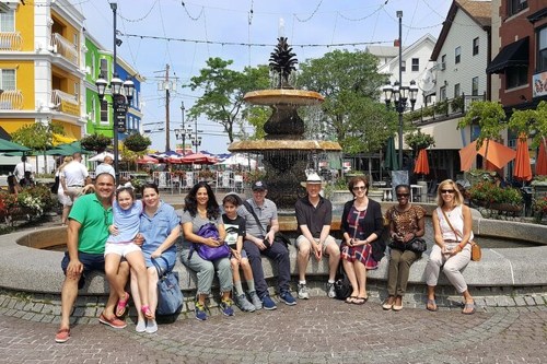 A diverse group of people sitting by a fountain in a vibrant outdoor plaza with colorful buildings and greenery.