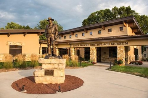 Statue of John Wayne in front of the John Wayne Birthplace Museum, surrounded by landscaping and a cloudy sky.