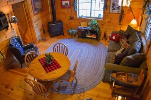 Cozy wooden cabin interior with a round table, chairs, a sofa, and a wood stove, surrounded by plants and decor.