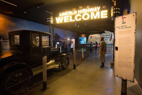 A vintage car in a museum exhibit featuring the Lincoln Highway, with a "Welcome" sign and informational displays.