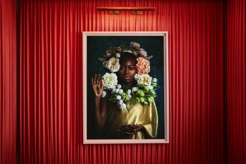 A portrait of a woman adorned with flowers, framed against a backdrop of red curtains. She holds her hand in a graceful pose.