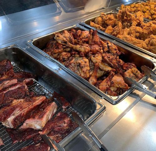 A buffet display featuring grilled meats, including ribs and chicken, in metal trays with tongs for serving.