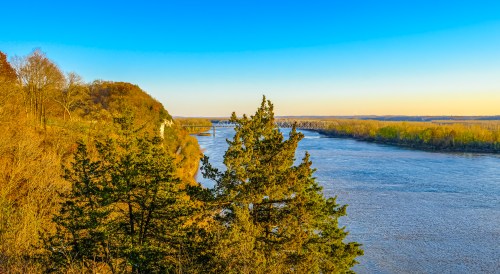 A serene river landscape with trees and hills under a clear blue sky during sunset.