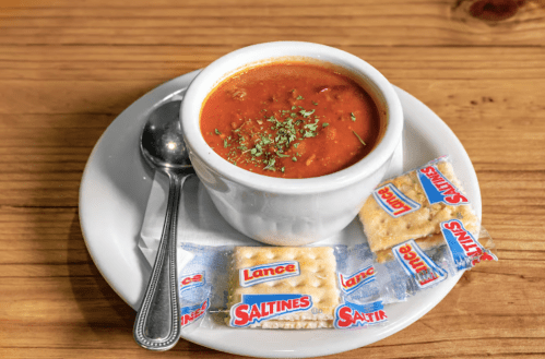 A bowl of tomato soup garnished with herbs, served with a spoon and Saltines on a white plate.