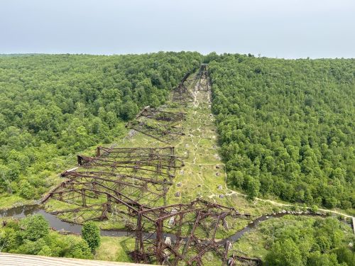 Aerial view of a rusted, abandoned structure winding through a lush green landscape with trees and a small stream.