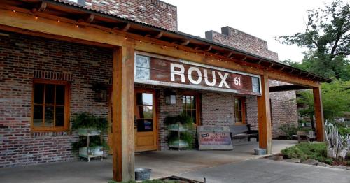 Exterior of a rustic brick building with a wooden awning, featuring the sign "Roux 61" and potted plants.