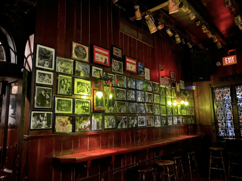 A dimly lit bar wall adorned with numerous black-and-white photos and framed memorabilia, with a wooden counter below.