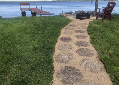 A stone pathway leads to a dock by a lake, surrounded by green grass and a fire pit nearby.