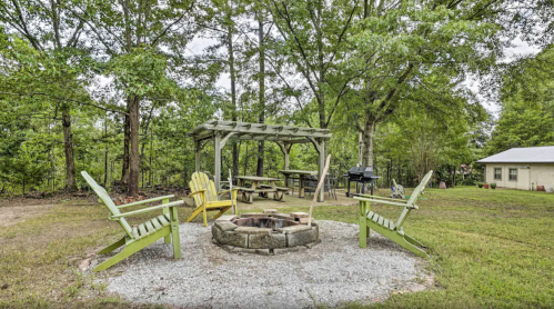 A cozy outdoor space with a fire pit surrounded by chairs, picnic tables, and trees in a green setting.