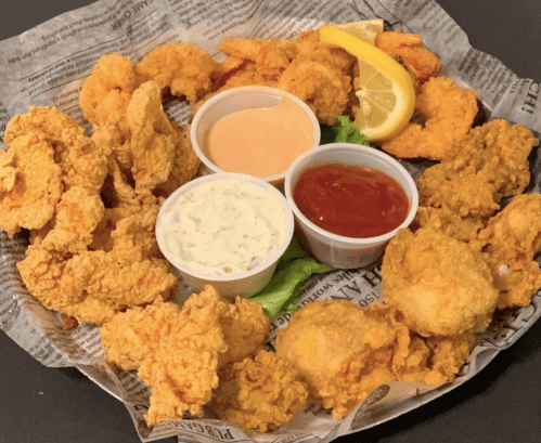 A platter of fried shrimp and chicken, served with dipping sauces and a lemon wedge, on a bed of newspaper.