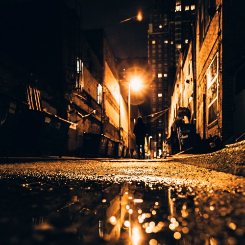 A dimly lit alley at night, with a figure walking and reflections in puddles on the wet ground.