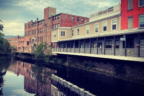 Historic buildings line a calm river, reflecting their colors on the water's surface under a cloudy sky.