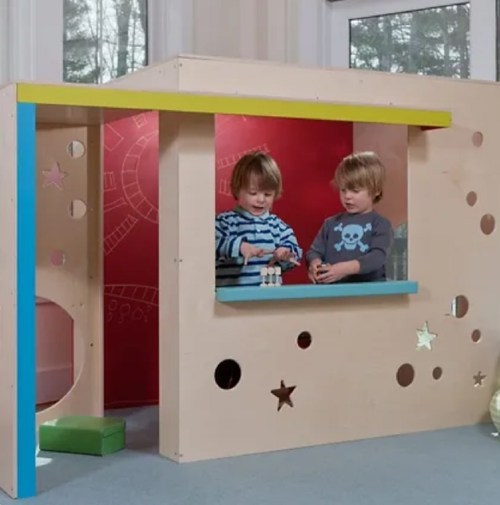 Two children play at a colorful wooden playhouse with a window, surrounded by playful cutouts and designs.