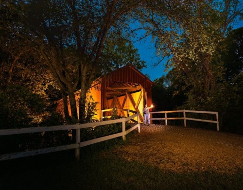 A covered bridge illuminated at night, surrounded by trees and a gravel path, with a person holding a lantern.