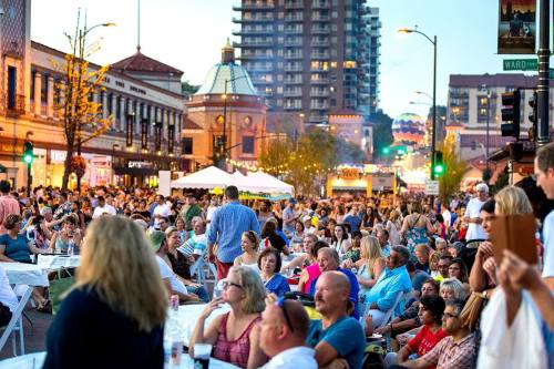 A large crowd gathers on a lively street during an outdoor event, with tents and buildings in the background.