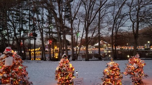 Snow-covered park with decorated Christmas trees and festive lights reflecting on a nearby lake at dusk.