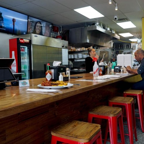 A cozy diner interior with a wooden counter, red stools, and a staff member serving customers in the background.