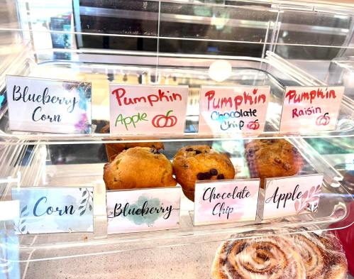 A display case with various muffins labeled: Blueberry, Pumpkin Apple, Pumpkin Chocolate Chip, Pumpkin Raisin, Corn, Blueberry, Chocolate Chip, and Apple.