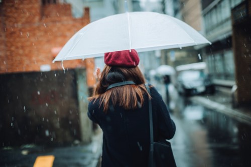 A person with a red beret walks in the rain, holding a clear umbrella on a city street.