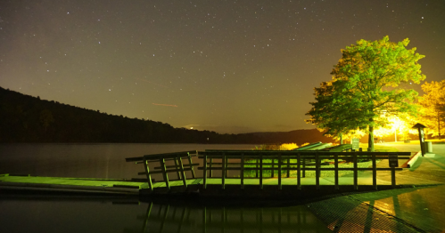 A serene lakeside scene at night, featuring a wooden dock, illuminated trees, and a starry sky.