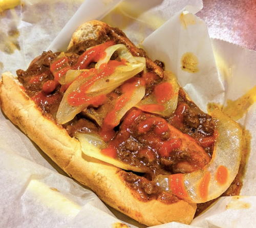 A loaded sandwich with beef, sautéed onions, and ketchup on a toasted bun, served in a paper-lined basket.